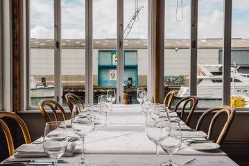 A table set up with white table cloth, six chairs, wine and water glasses, utensils and side plates.