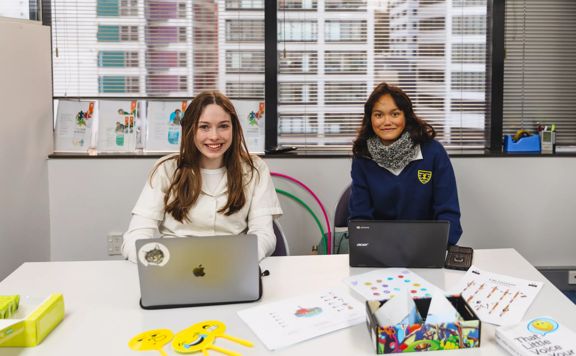 Two smiling students working on their laptops at a table.