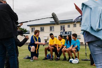 Behind the scenes on the set of  ‘Extreme Cake Sports’, a Wellington-based reality TV show. Four contestants sit on a bench, a person is kneeling to their right holding a microphone towards them.