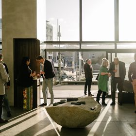 People mingle inside the James Blackie Gallery as sunlight spills into the space from outside.