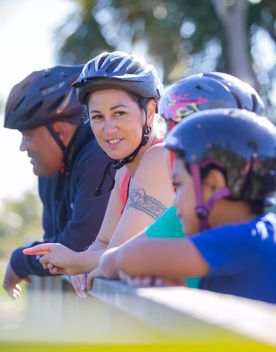 Four people wearing bicycle helmets are standing, leaning against a wooden railing on a bridge along the Coast 35 trail in Queen Elizabeth Regional Park in the Kāpiti Coast.