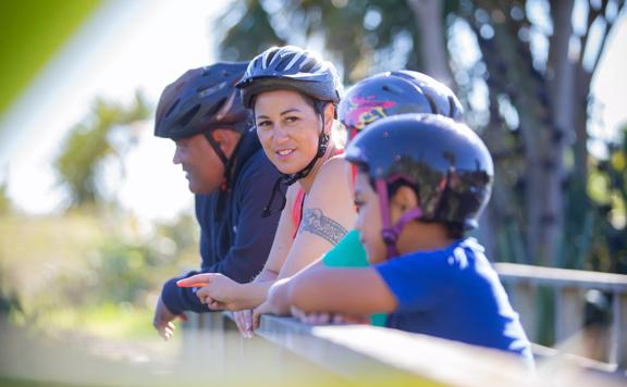 Four people wearing bicycle helmets are standing, leaning against a wooden railing on a bridge along the Coast 35 trail in Queen Elizabeth Regional Park in the Kāpiti Coast.