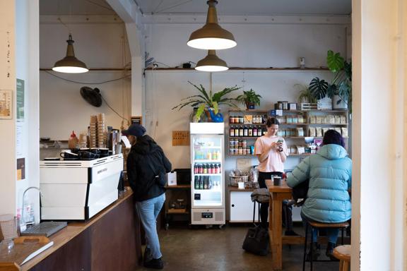 The interior of Peoples Coffee Constable  Street cafe located in Newtown, Wellington. There are three customers in the cafe, the walls are white and there is wood furniture and four green potted house plants. 