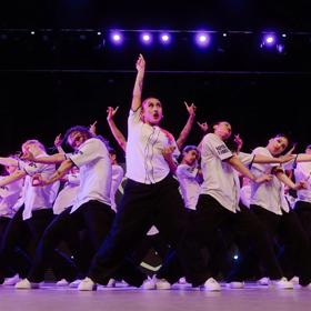 A large group of twenty-plus dancers from The Royal Family Dance Crew stand in a triangular group, with a main dancer in the front holding their hands up, and everyone else to the side. They all wear white shirts and black pants.