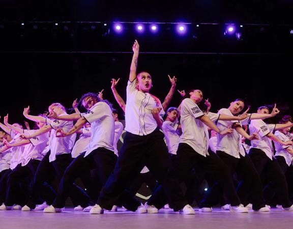 A large group of twenty-plus dancers from The Royal Family Dance Crew stand in a triangular group, with a main dancer in the front holding their hands up, and everyone else to the side. They all wear white shirts and black pants.