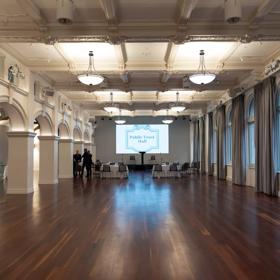 Very far away photo of conference room with hard wood floors, 4 tables each with 4 chairs, facing towards a projector in the centre of the room and podium.