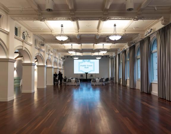 Very far away photo of conference room with hard wood floors, 4 tables each with 4 chairs, facing towards a projector in the centre of the room and podium.