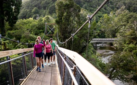 People walking along one of the two swing bridges over the river amongst trees in Kaitoke Regional Park.