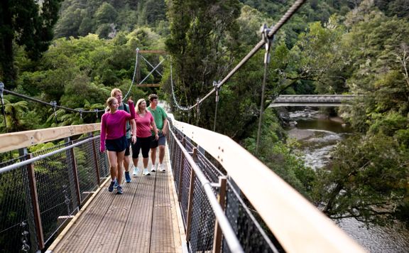 People walking along one of the two swing bridges over the river amongst trees in Kaitoke Regional Park.