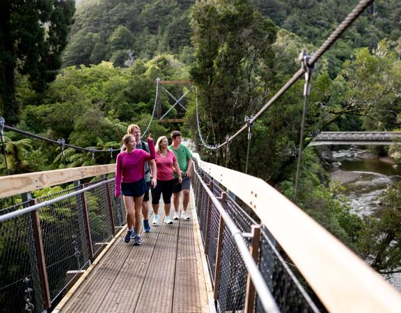 People walking along one of the two swing bridges over the river amongst trees in Kaitoke Regional Park.