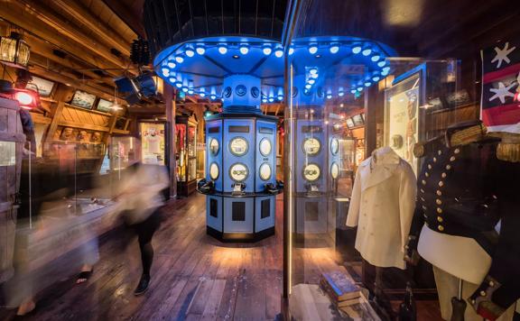 Inside a room at the Wellington Museum with wood flooring, wood-paneled walls and ceiling, two mannequin busts dressed in nautical jackets and three rotary phones displayed on a blue octagonal pillar in the middle of the room.
