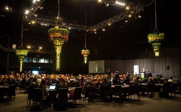 Large conference room with hundreds of people sitting at circular tables and dark lighting, with colourful chandeliers hanging above. Taken from standing perspective.