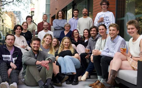 A group of 20 people, a few with drinks in their hands, gathered together and smiling on a balcony in an urban setting.