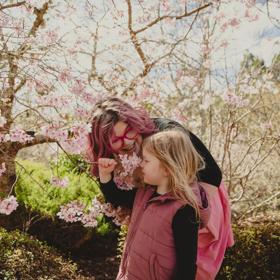 Parent and child at Blossom Valley. Parent smiles down at child, whilst child sniffs blossom flowers.