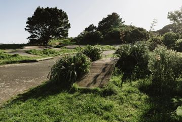 Mountain bike skills area with a wooden jump track surrounded by grass and a concrete pump track in the background.
