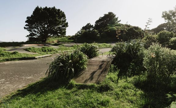 Mountain bike skills area with a wooden jump track surrounded by grass and a concrete pump track in the background.