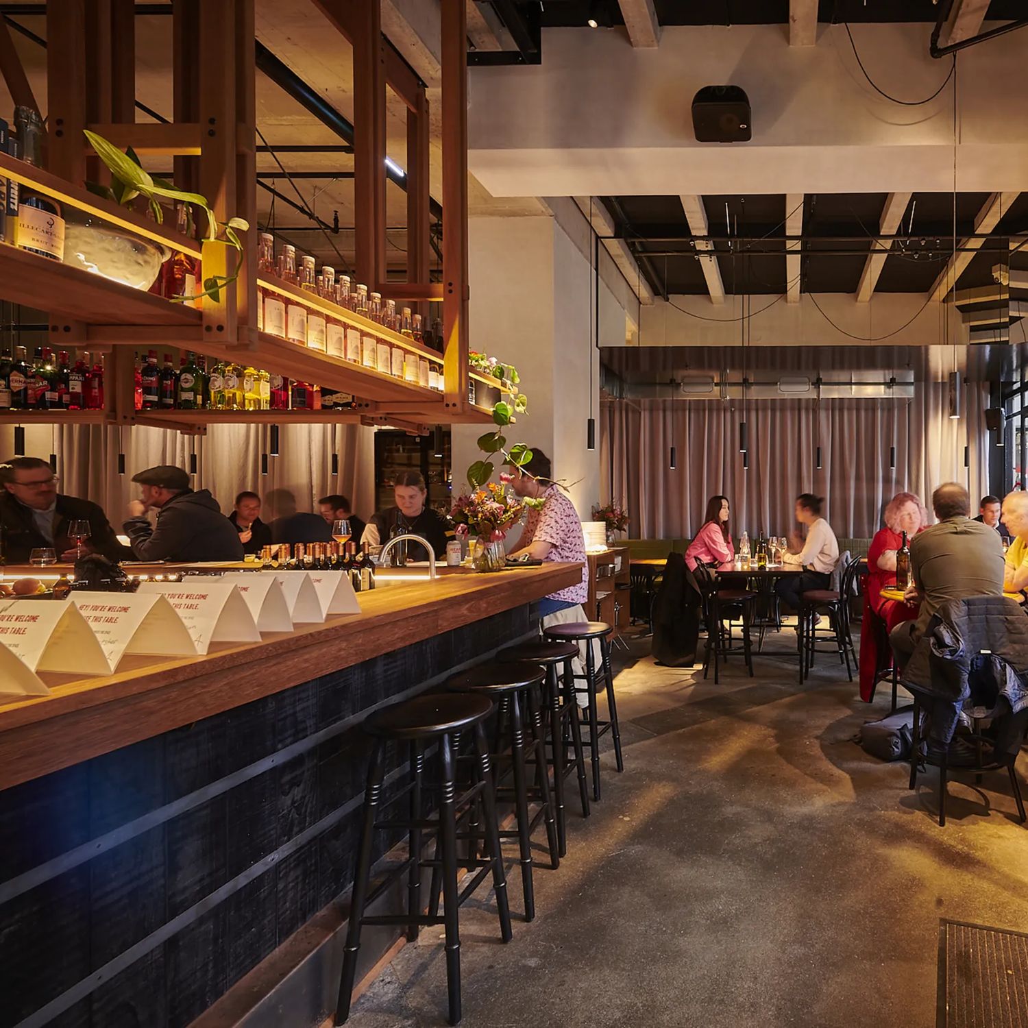 A wide interior shot of Astoria. A large black wooden bar is on the left, and people sit at tables on the right—the lighting is dim and ambient.