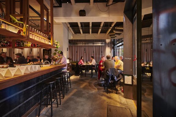 A wide interior shot of Astoria. A large black wooden bar is on the left, and people sit at tables on the right—the lighting is dim and ambient.