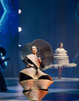 A model wears an avant-garde dress made of a golden ribbon with a black and red pattern wrapped around their body, holding a fishnet umbrella.
