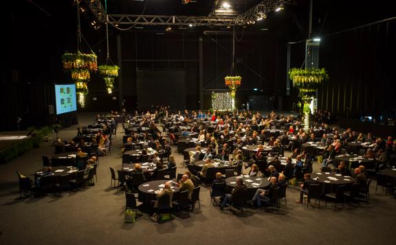 Large conference room with hundreds of people sitting at circular tables and dark lighting, with colourful chandeliers hanging above. Taken from above.