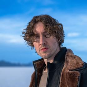 Australian pop singer-songwriter, Dean Lewis, wearing a black jacket looks into the camera with a blue sky in the background. 