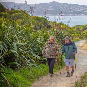 Two people walking along Te Onepoto trail in Porirua. Walkng canes are assisting both. The harbour and hills of Porirua are in the background.