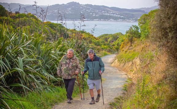 Two people walking along Te Onepoto trail in Porirua. Walkng canes are assisting both. The harbour and hills of Porirua are in the background.