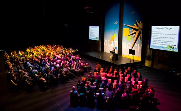 A presentation at the TSB Arena located on Queens Wharf in Wellington Central. There is a person standing behind a podium on a small stage while an audience sits an listens.