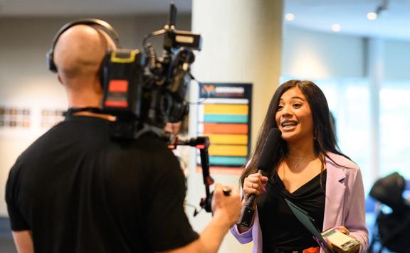 A cameraman shoots a reporter wearing a pink blazer.