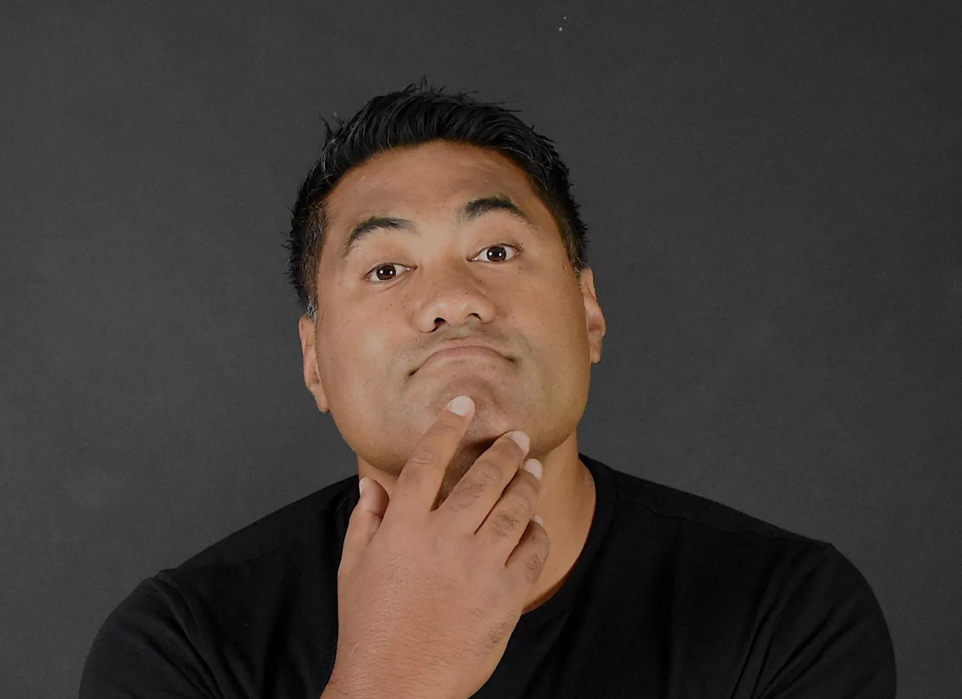 Stand-up comedian, Li'i Alaimoana, wears a black T-shirt, poses in front of a grey background with his right hand touching his chin.