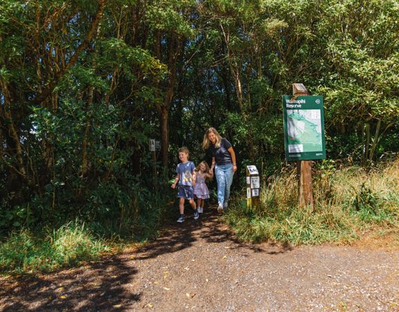 An adult and two children exit a forest trail next to a sign post with a map.