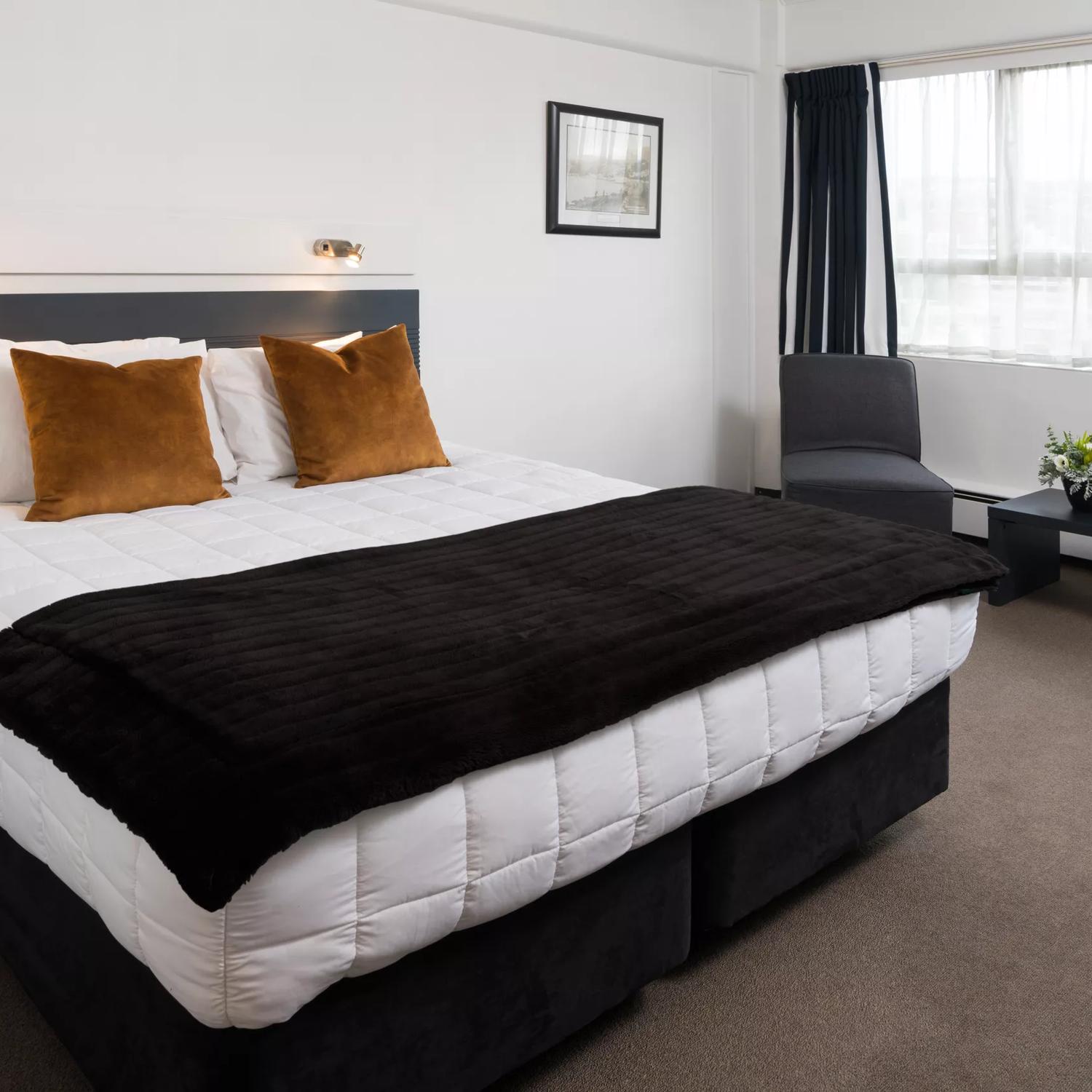 A super king bed within a hotel room at Bay Plaza Hotel, with two chairs and a small table to the right, in front of a window.