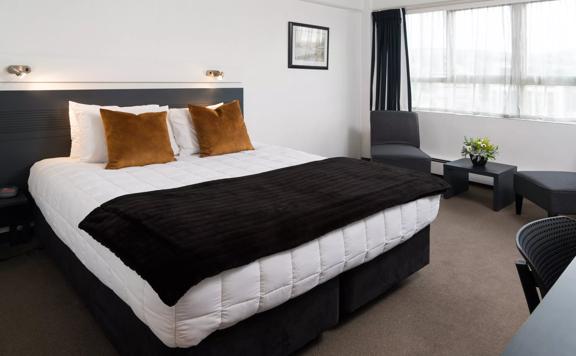 A super king bed within a hotel room at Bay Plaza Hotel, with two chairs and a small table to the right, in front of a window.