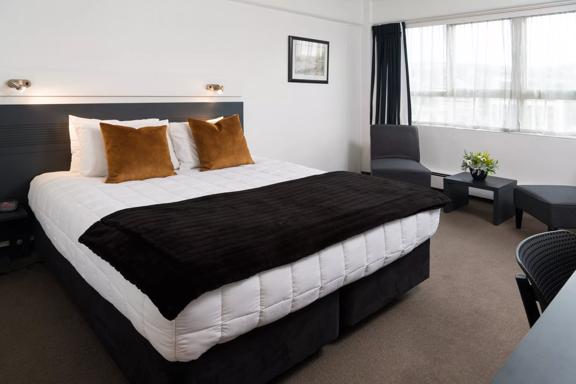 A super king bed within a hotel room at Bay Plaza Hotel, with two chairs and a small table to the right, in front of a window.