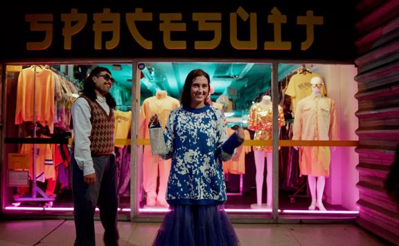 2 people standing outside the 'SPACESUIT' thrift store on Cuba Street at night time, with neon lights around them. One character is holding a purse, wearing an oversized blue jumper and blue skirt, and the other wears a vest and glasses. Mannequins sit in