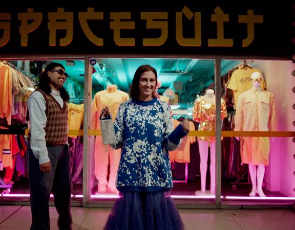 2 people standing outside the 'SPACESUIT' thrift store on Cuba Street at night time, with neon lights around them. One character is holding a purse, wearing an oversized blue jumper and blue skirt, and the other wears a vest and glasses. Mannequins sit in