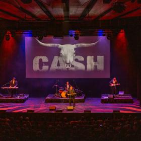 Johnny Cash & The Outlaws performing on stage under dramatic blue and red lighting, with a packed audience in view. A large projection screen behind them displays a bull skull and the word 'CASH.'