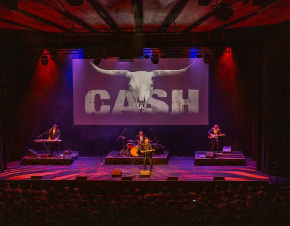 Johnny Cash & The Outlaws performing on stage under dramatic blue and red lighting, with a packed audience in view. A large projection screen behind them displays a bull skull and the word 'CASH.'