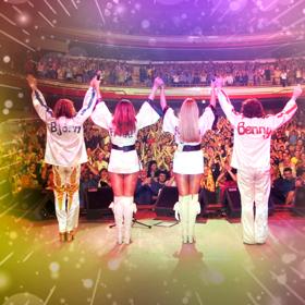 Four people in white disco outfits take a bow on stage in front of an audience.