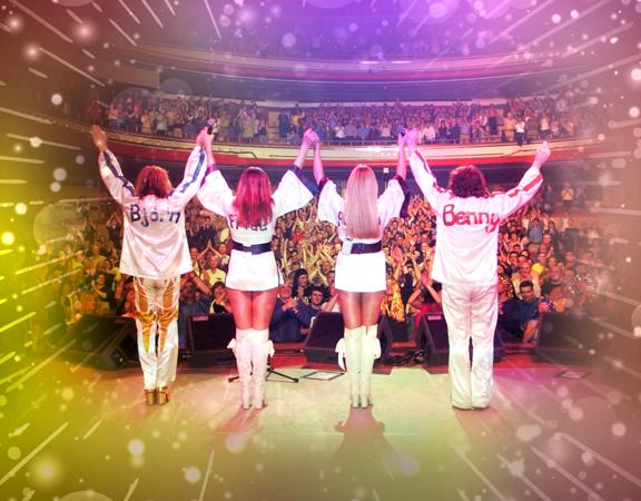 Four people in white disco outfits take a bow on stage in front of an audience.