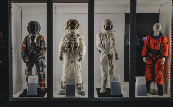 Four life-sized mannequins wearing spacesuits behind a glass enclosure. The one on the left is wearing a dark grey and orange suit, the two in the middle are wearing white suits, and the one on the right is wearing an orange suit with no helmet.