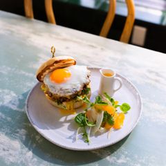 A breakfast sandwich topped with a fried egg is served on a plate with a small salad and a jug of sauce on a light-colored table.