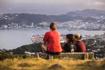 Three people sit on a bench looking at the view from the Skyline Walkway, a hiking trail in Wellington, New Zealand.