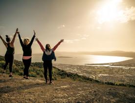 Three people are standing with their arms raised facing the view of the harbour in the background at Te Whiti Riser, a hiking trail in Lower Hutt Wellington.