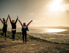 Three people are standing with their arms raised facing the view of the harbour in the background at Te Whiti Riser, a hiking trail in Lower Hutt Wellington.