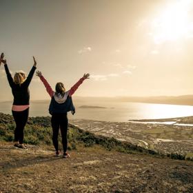 Three people are standing with their arms raised facing the view of the harbour in the background at Te Whiti Riser, a hiking trail in Lower Hutt Wellington.