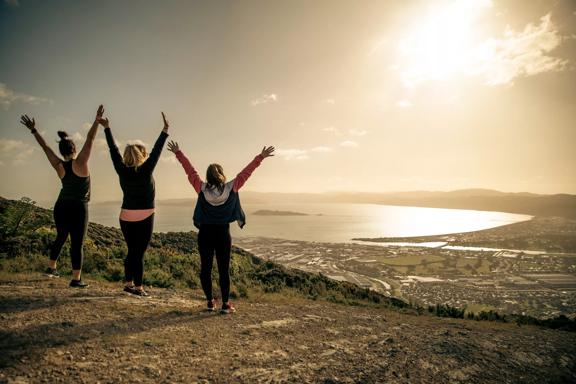 Three people are standing with their arms raised facing the view of the harbour in the background at Te Whiti Riser, a hiking trail in Lower Hutt Wellington.