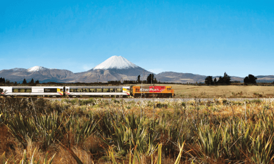 A KiwiRail train on route with blue skies above and a mountain range in the background.