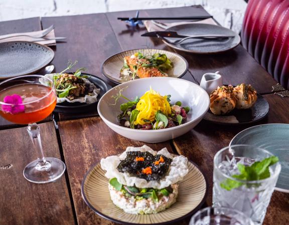 Five Asian-fusion dishes, a pink cocktail, dishes and utensils on a table at a restaurant.