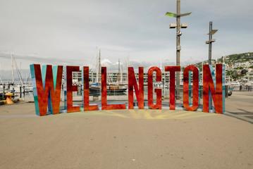 The Well_ngton Sign is located at the Wellington Harbourfront Walk, with a metallic red facade reflecting light onto the cement in the foreground.